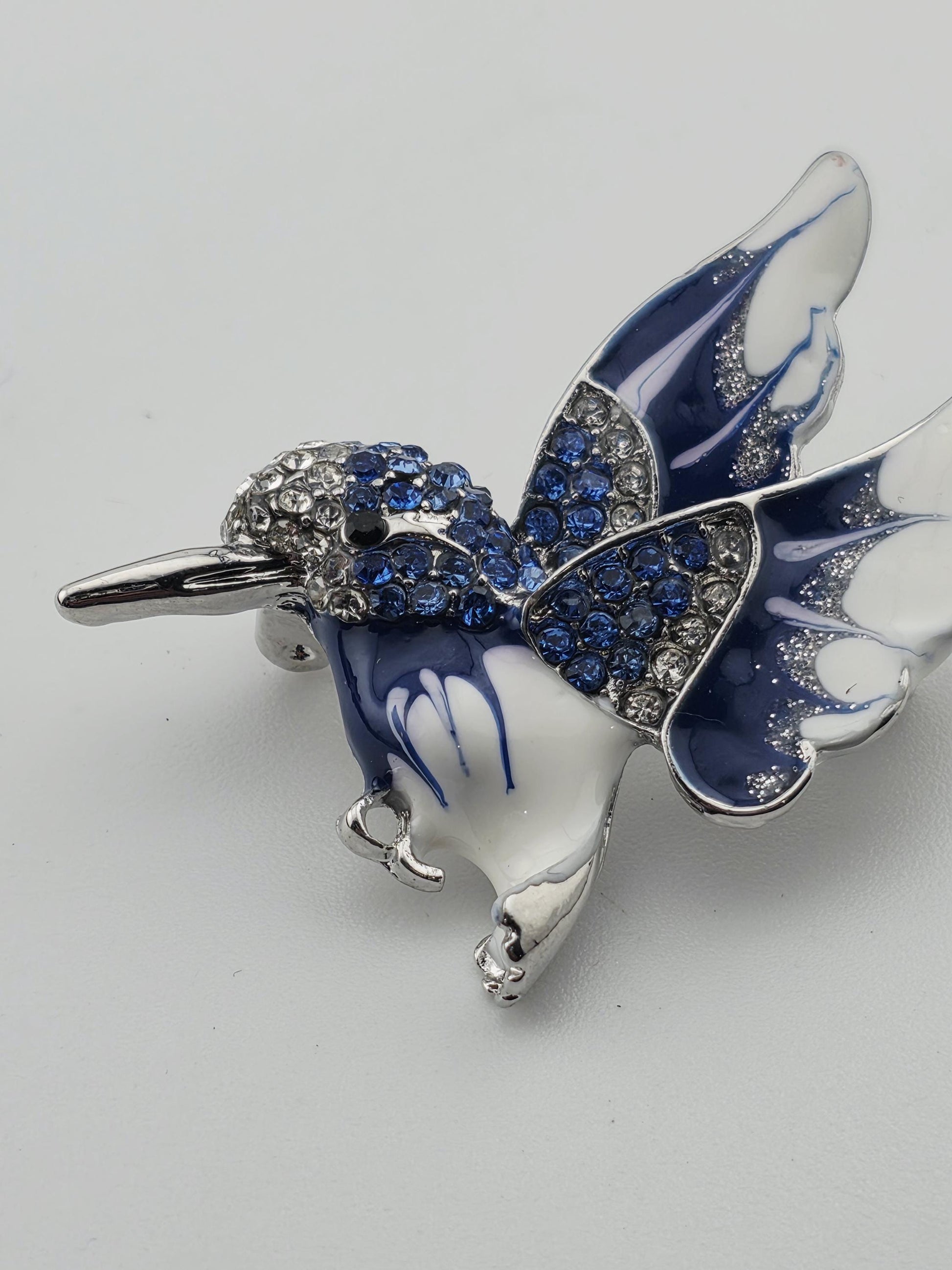 A decorative brooch in the shape of a bird, with blue and white colors and sparkling rhinestones or crystals on its wings and body.