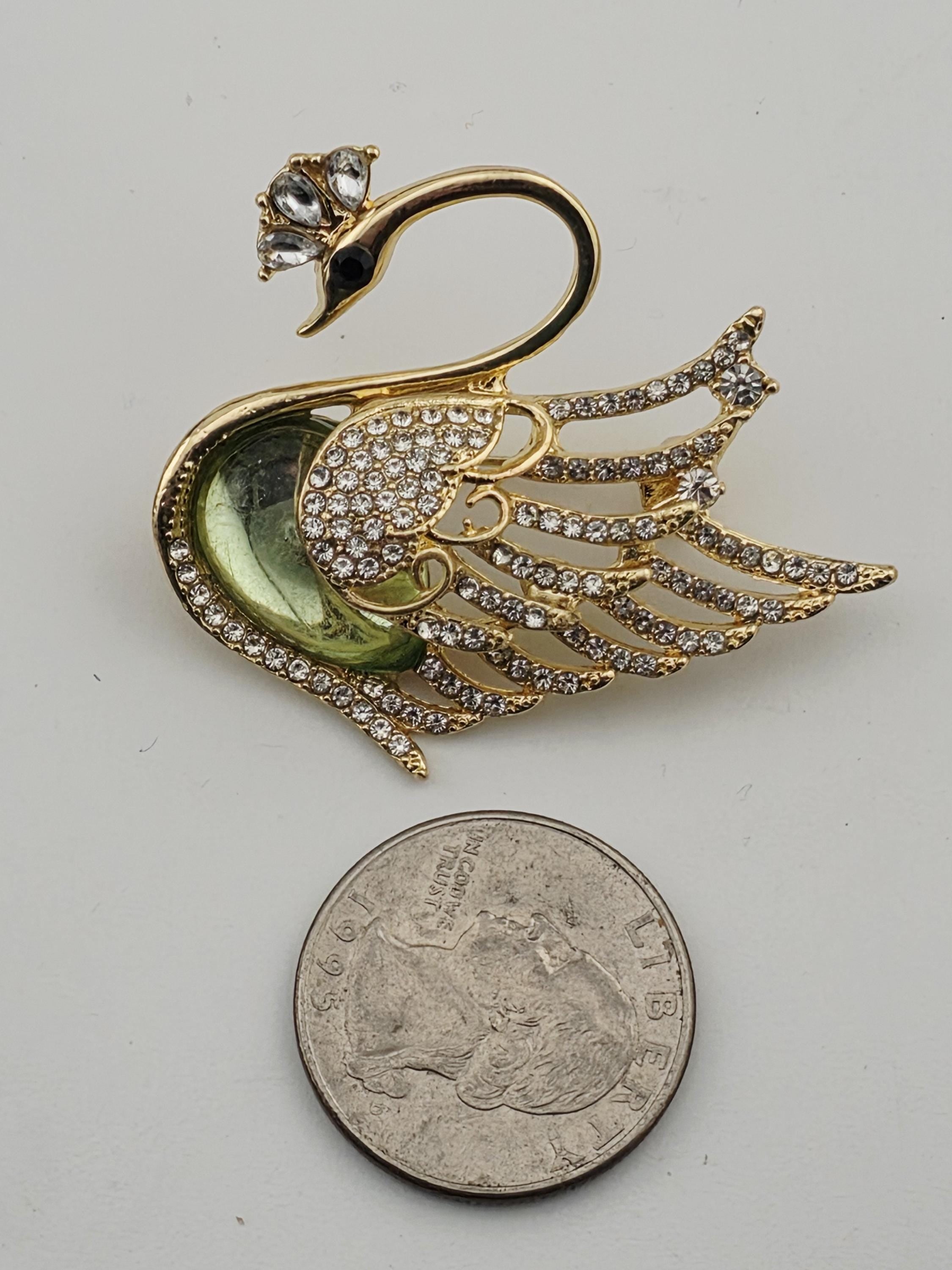 A gold-colored swan brooch with a green gemstone and sparkling rhinestones is placed next to a quarter coin.