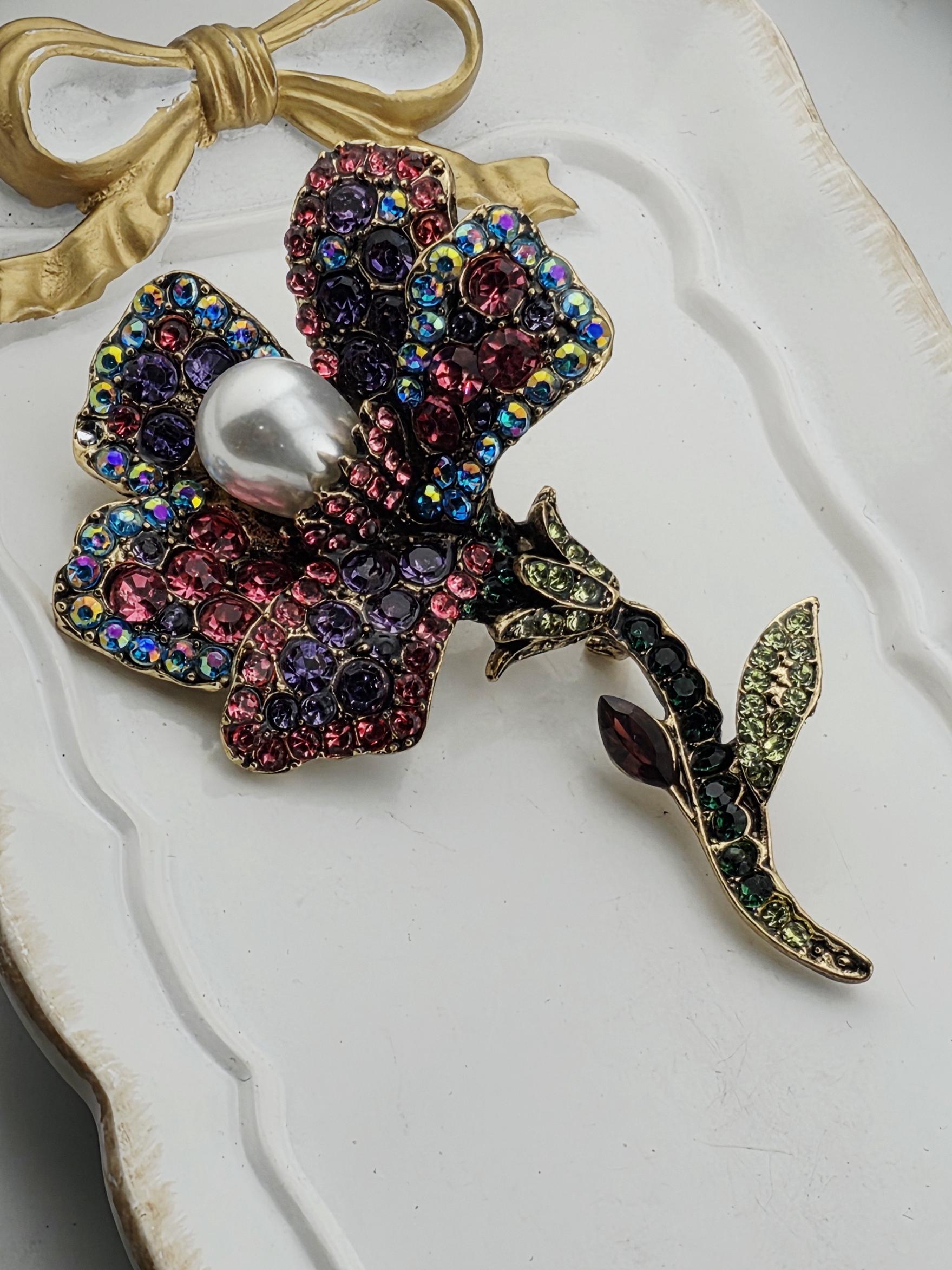 A decorative brooch with a flower-like design, featuring a variety of colorful gemstones and a large pearl at its center.