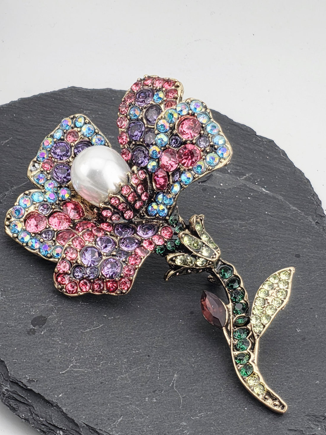 A decorative brooch with a flower-like design, featuring a variety of colorful gemstones and a large pearl at its center.