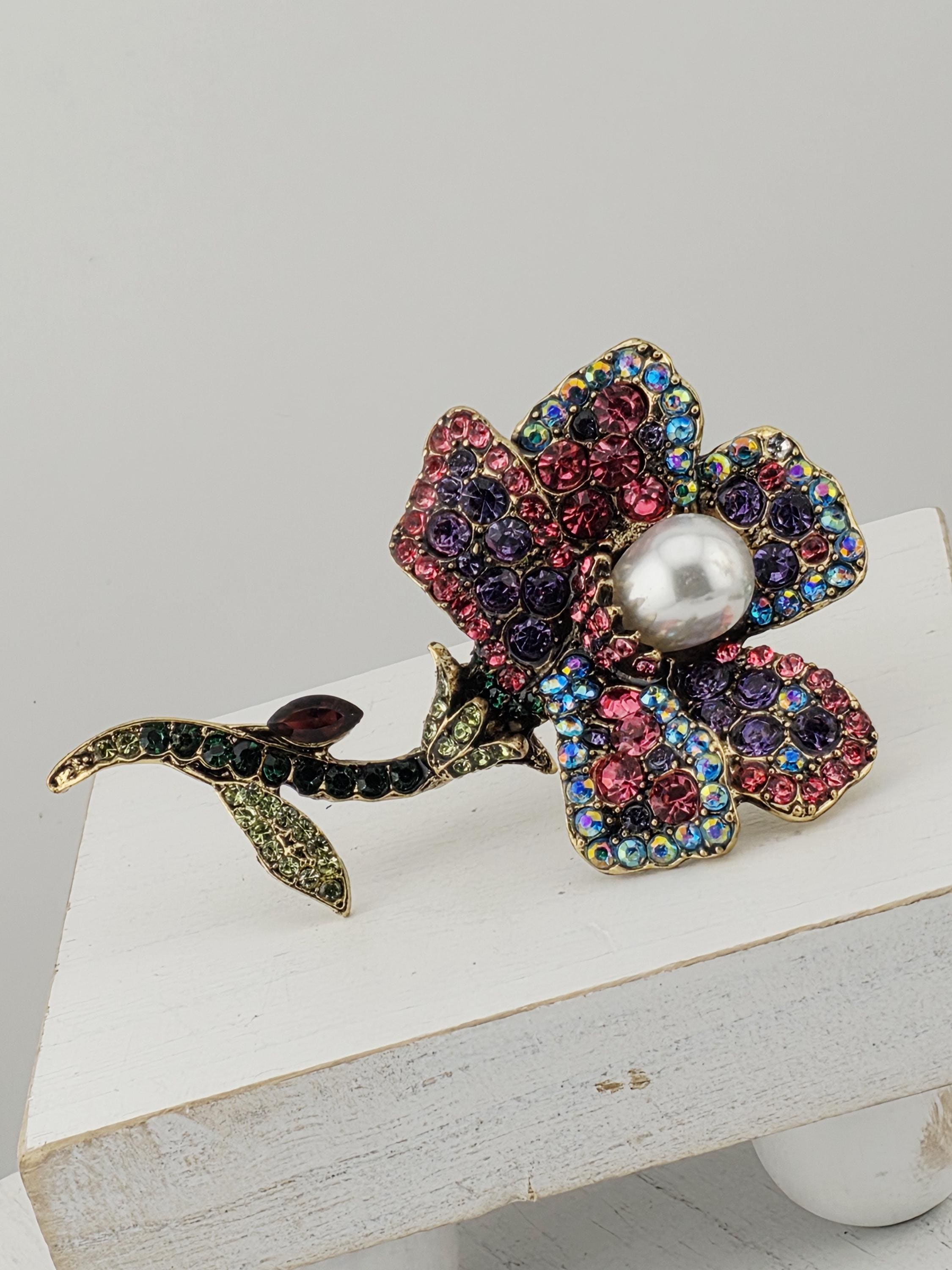A decorative brooch with a flower-shaped design, featuring a variety of colorful gemstones and a large pearl at the center.