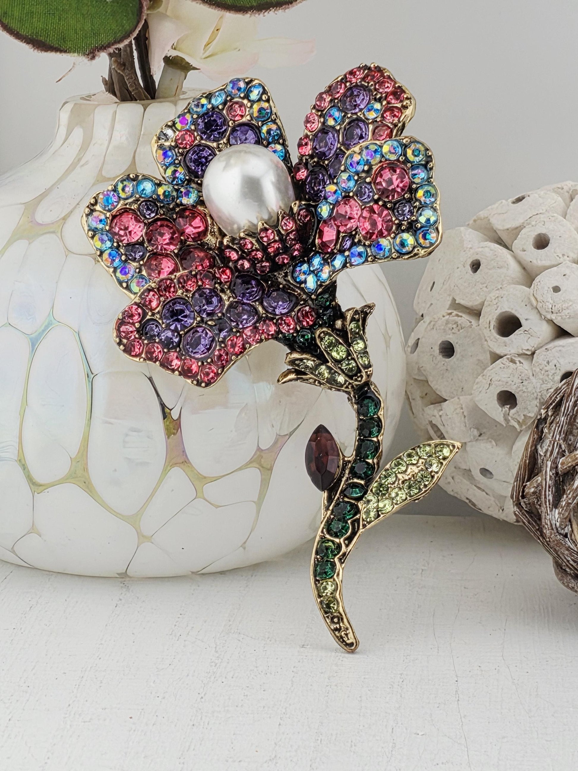 A decorative brooch with a flower-like design, adorned with colorful gemstones and a pearl at its center, is displayed against a white background.