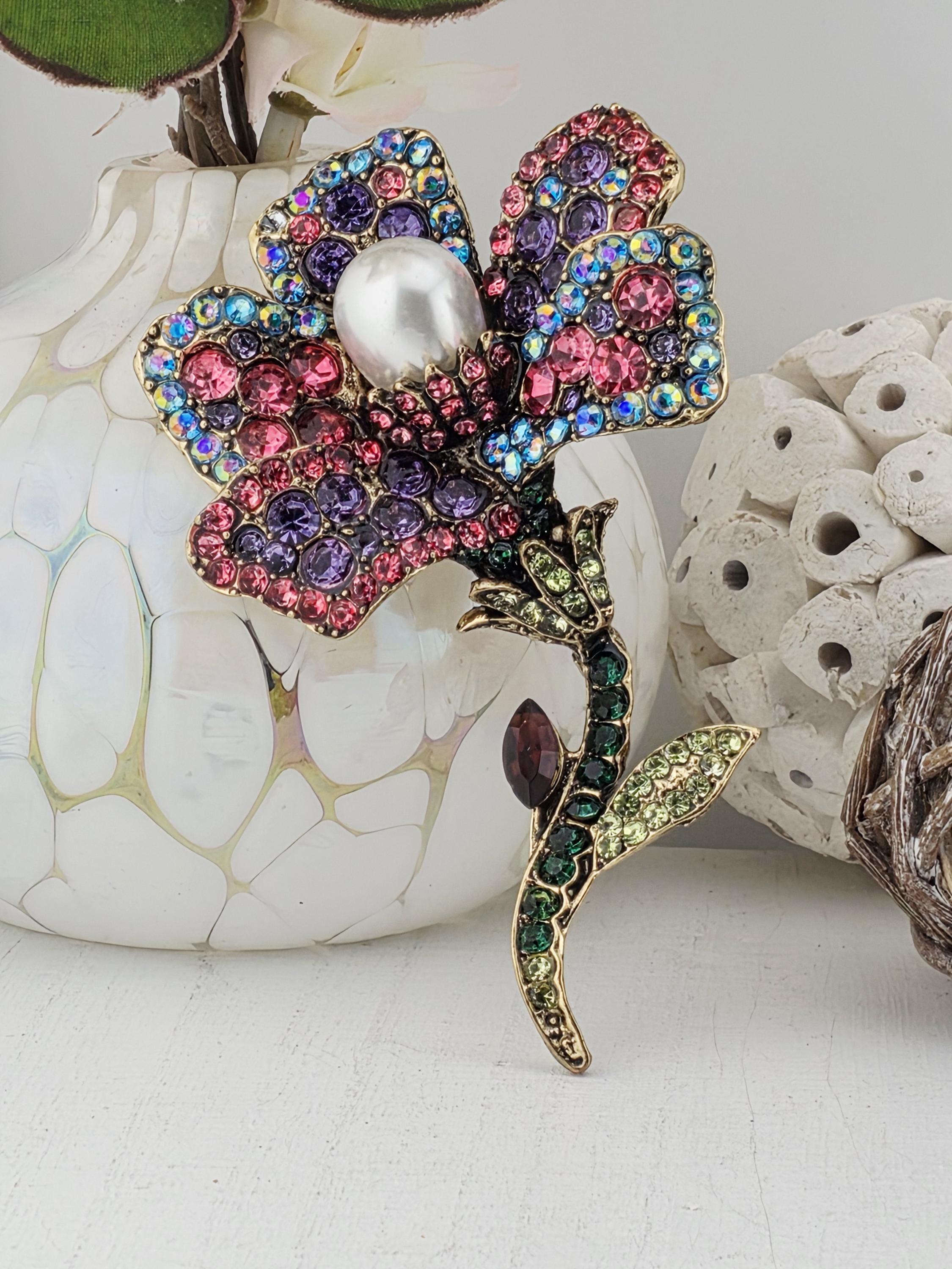 A decorative brooch with a flower-like design, adorned with colorful gemstones and a pearl at its center, is displayed against a white background.