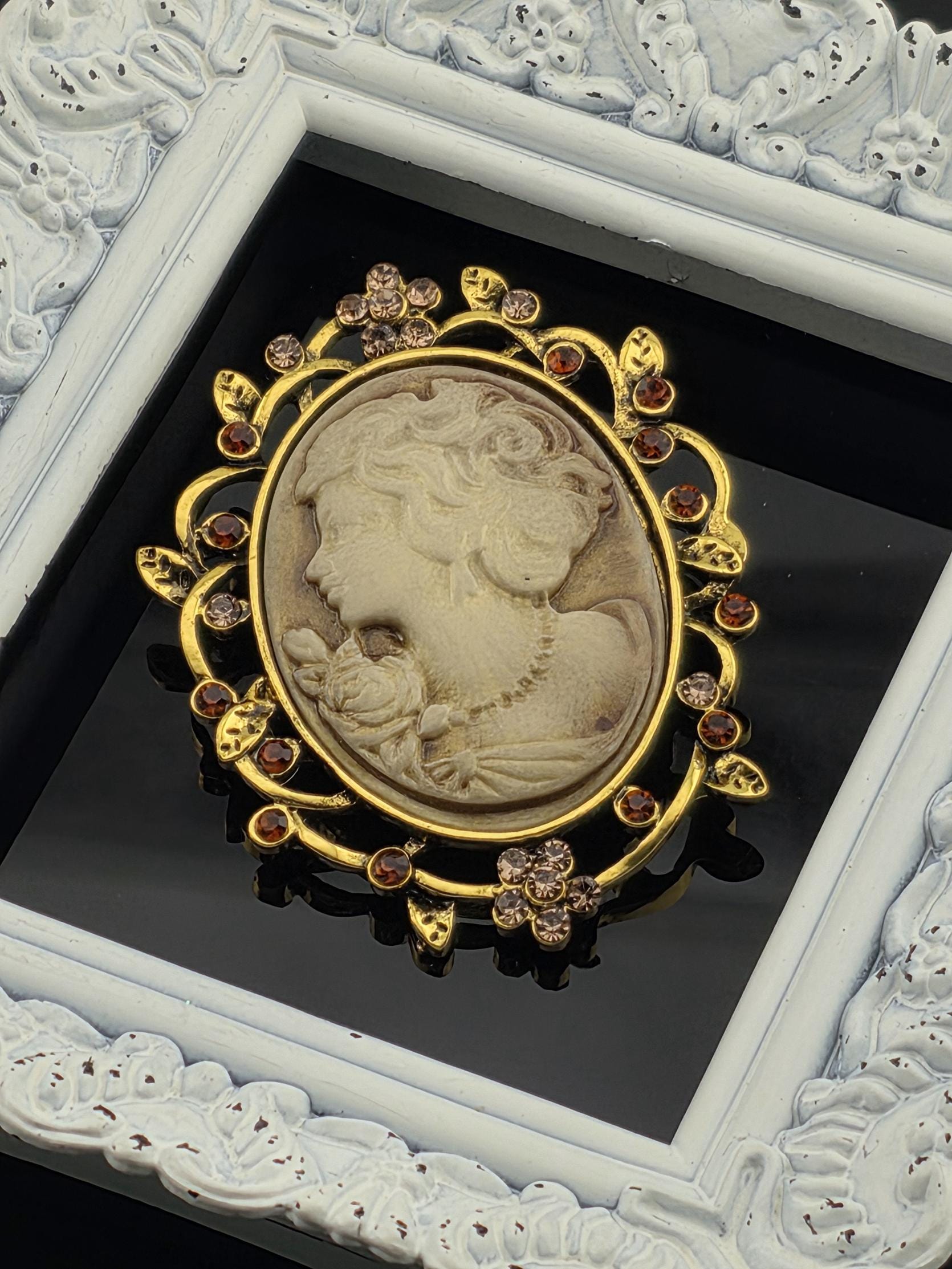 A decorative oval brooch with a carved face and floral elements, surrounded by a gold frame with a white border.