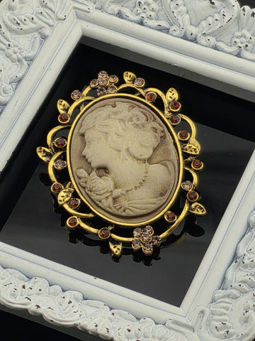 A decorative oval brooch with a carved face and floral elements, surrounded by a gold frame with a white border.