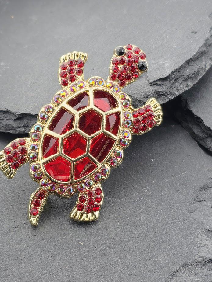 A decorative red and gold turtle brooch with a red gemstone shell and a gold shell, placed on a gray surface.