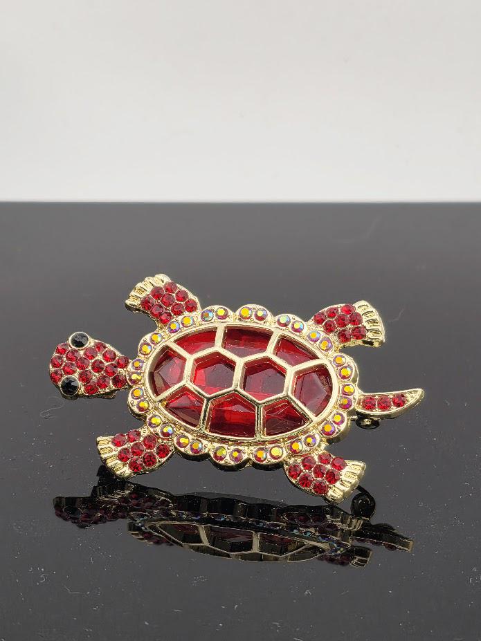 A decorative red and gold turtle brooch with rhinestones on its shell and legs, displayed on a black surface.