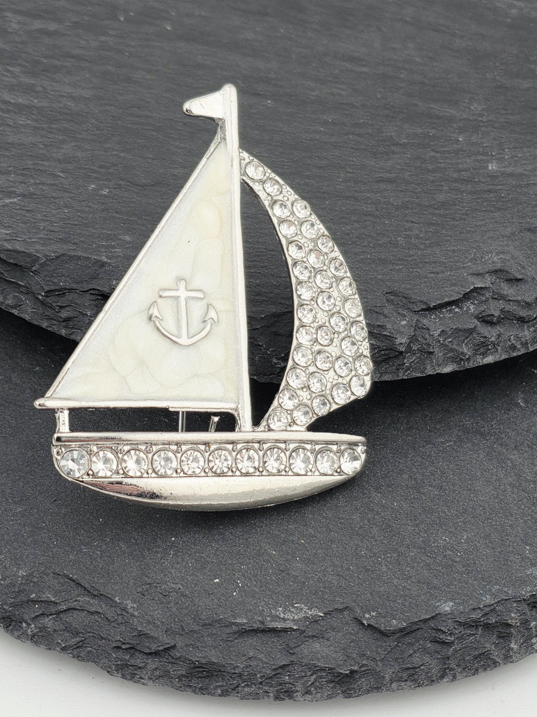 A silver and white brooch with a gold ribbon and a small anchor symbol is displayed on a white surface.