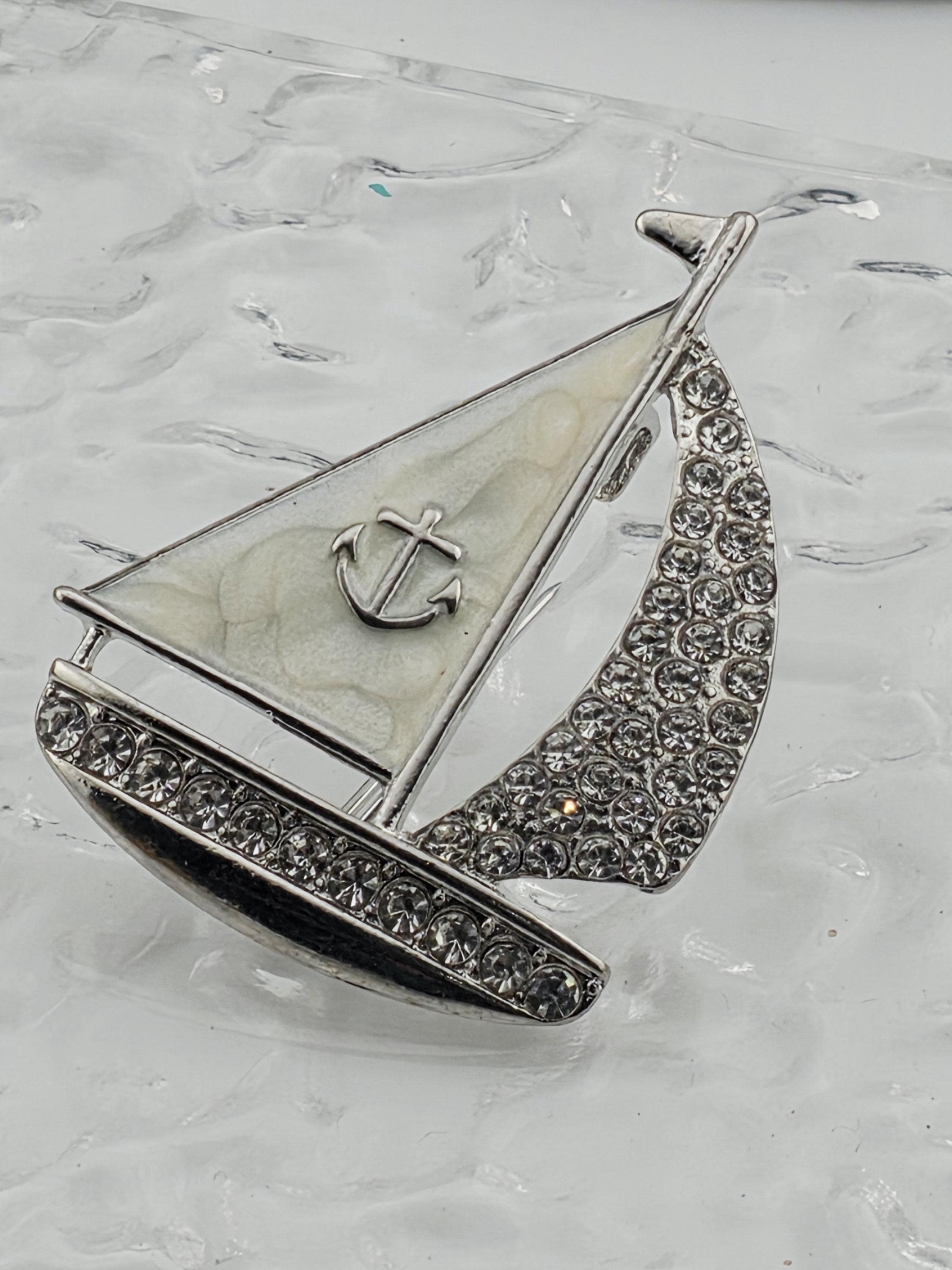 A silver-colored brooch with a boat-shaped design and a small anchor symbol, set against a white background.
