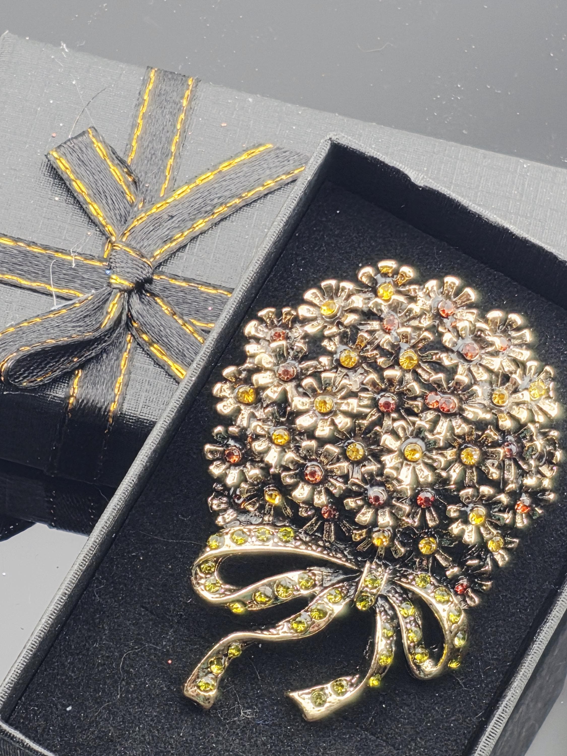 A decorative brooch with a floral design and yellow gemstones is displayed in a black box with a yellow ribbon bow.
