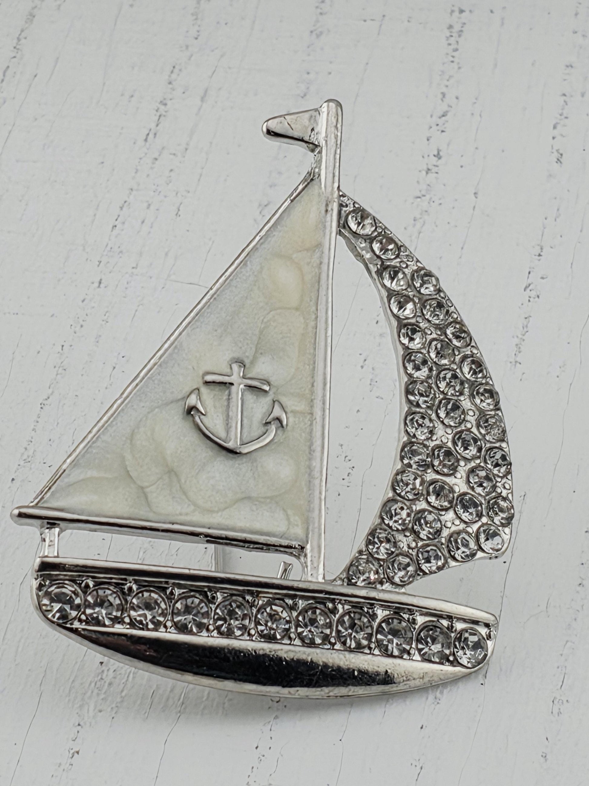 A silver-colored brooch with a white sailboat design and a cross symbol, adorned with small rhinestones or crystals.