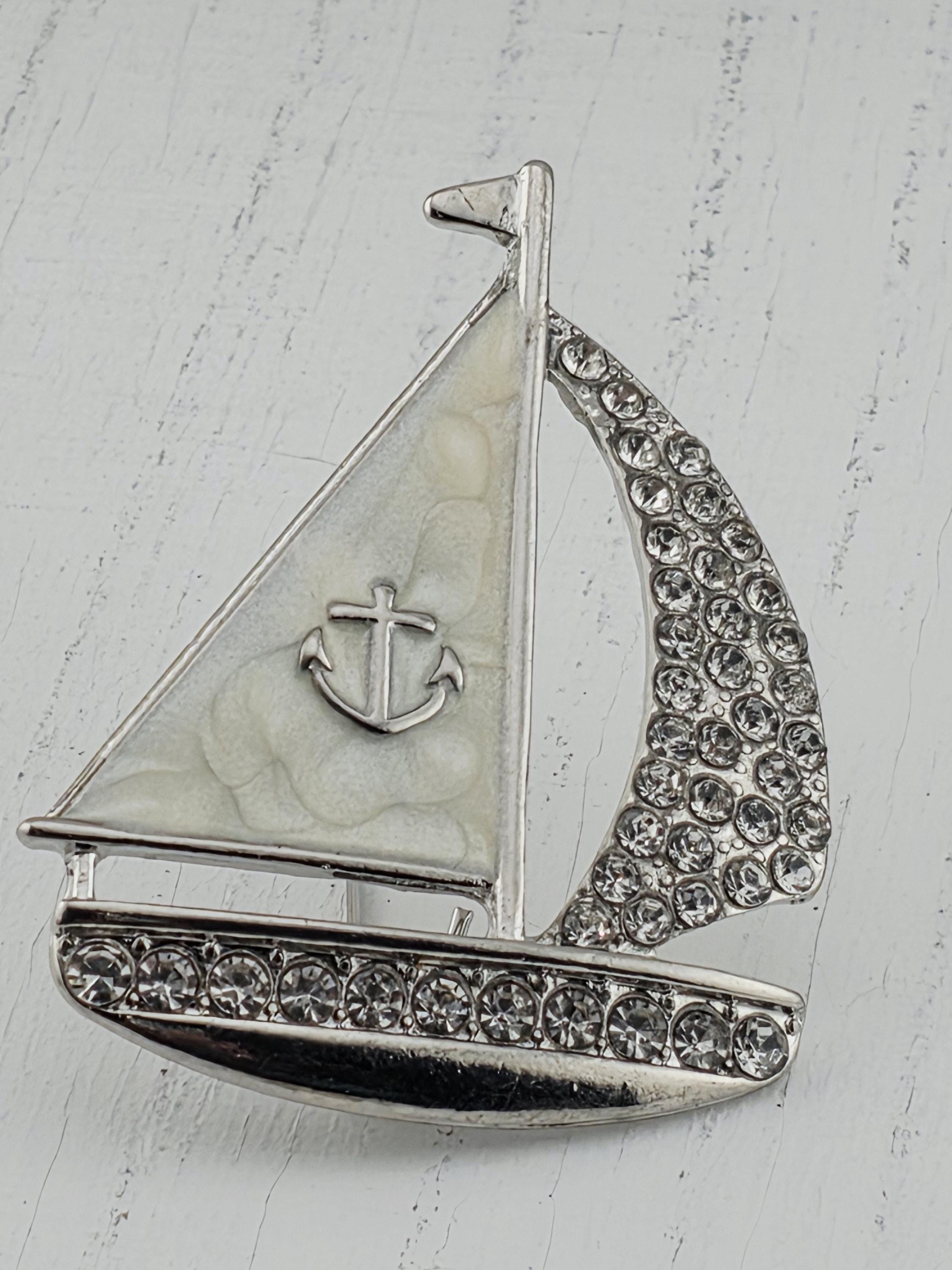 A silver-colored brooch with a white sailboat design and a cross symbol, adorned with small rhinestones or crystals.