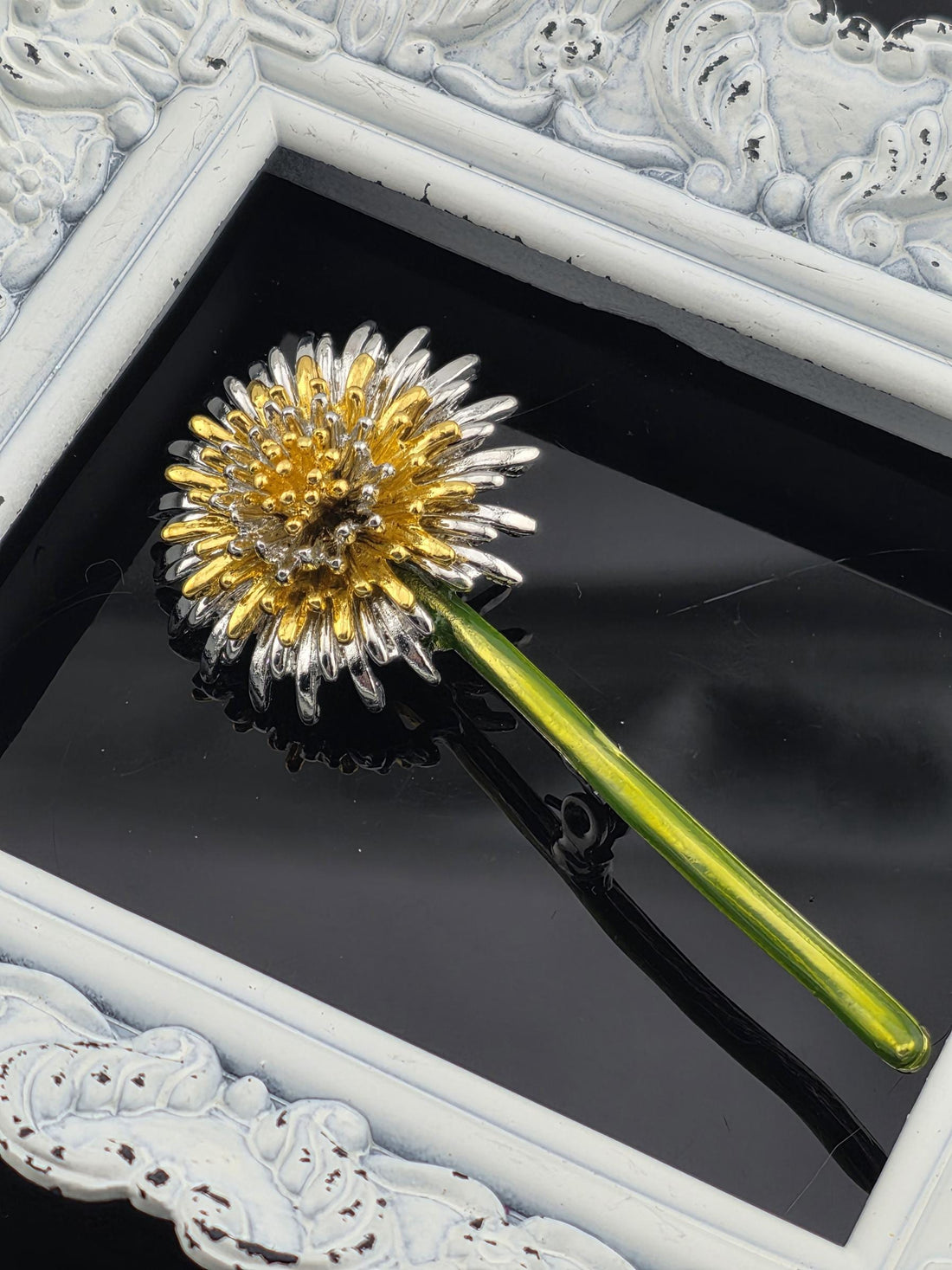 A decorative flower-shaped hair accessory with a green stem and gold and silver details, placed on a white surface.
