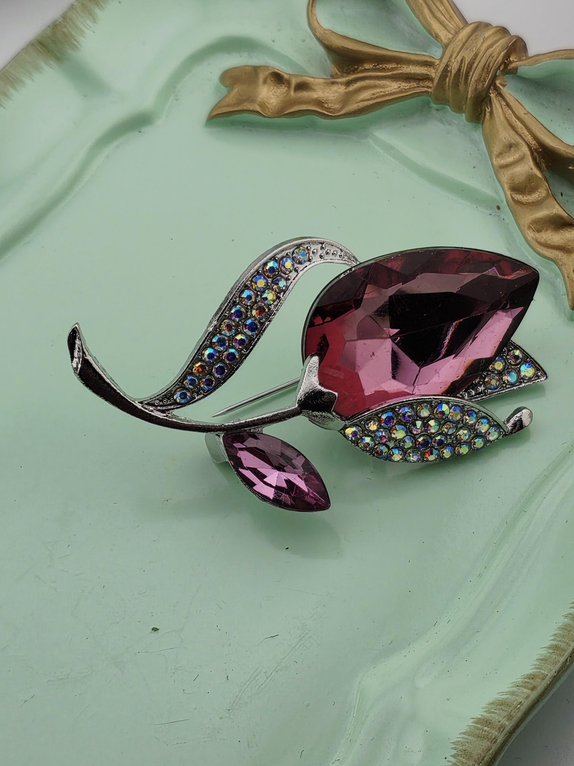 A decorative brooch with a large purple gemstone and smaller purple gemstones, set against a mint green background with a gold ribbon.