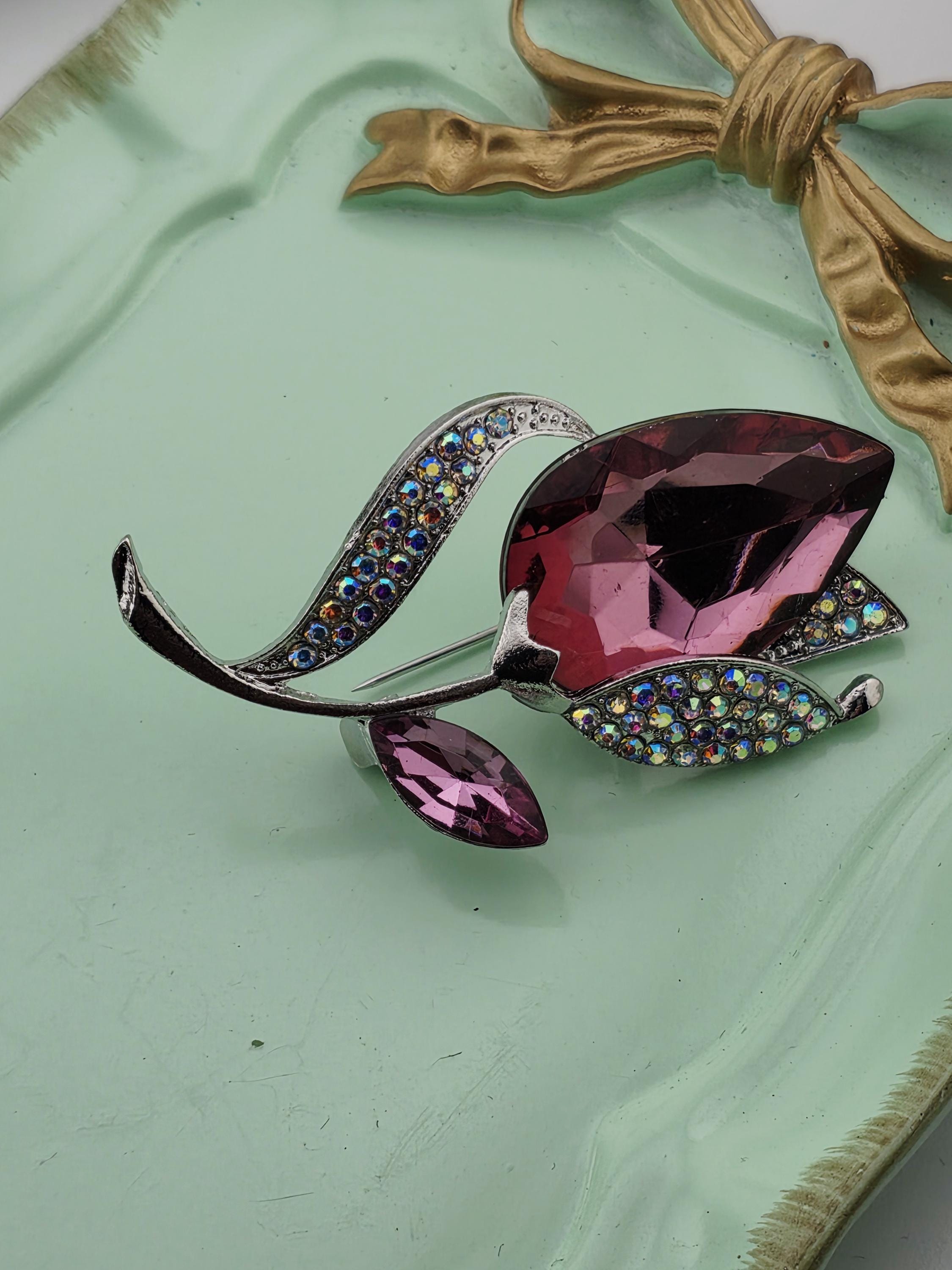 A decorative brooch with a large purple gemstone and smaller purple gemstones, set against a mint green background with a gold ribbon.