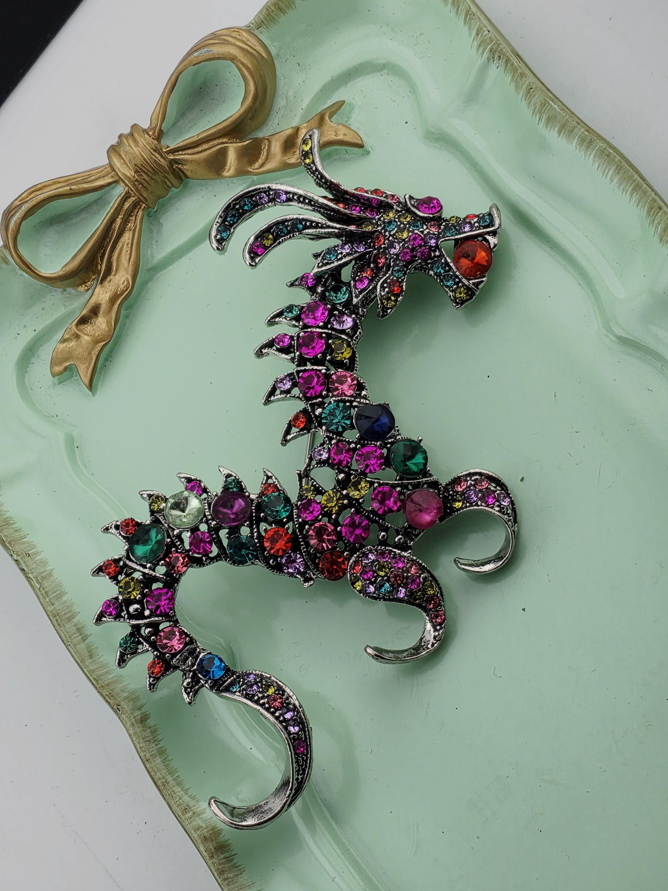 A decorative dragon-shaped brooch with a vibrant array of colorful gemstones, including red, green, blue, and purple stones, is displayed on a green tray.