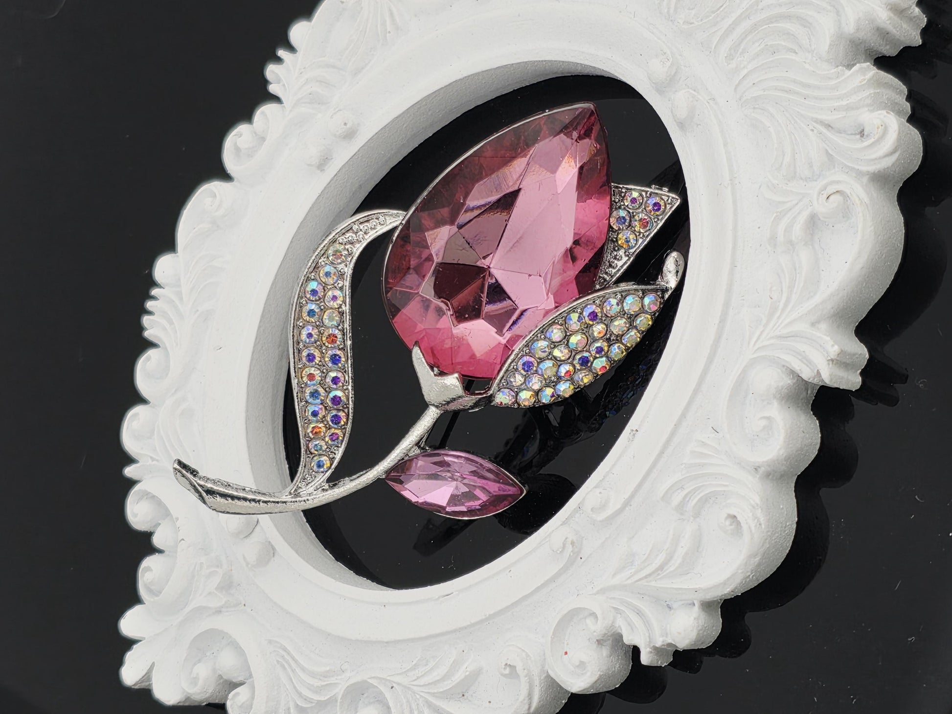 A pink gemstone brooch with a silver leaf design, placed within an ornate white frame against a black background.