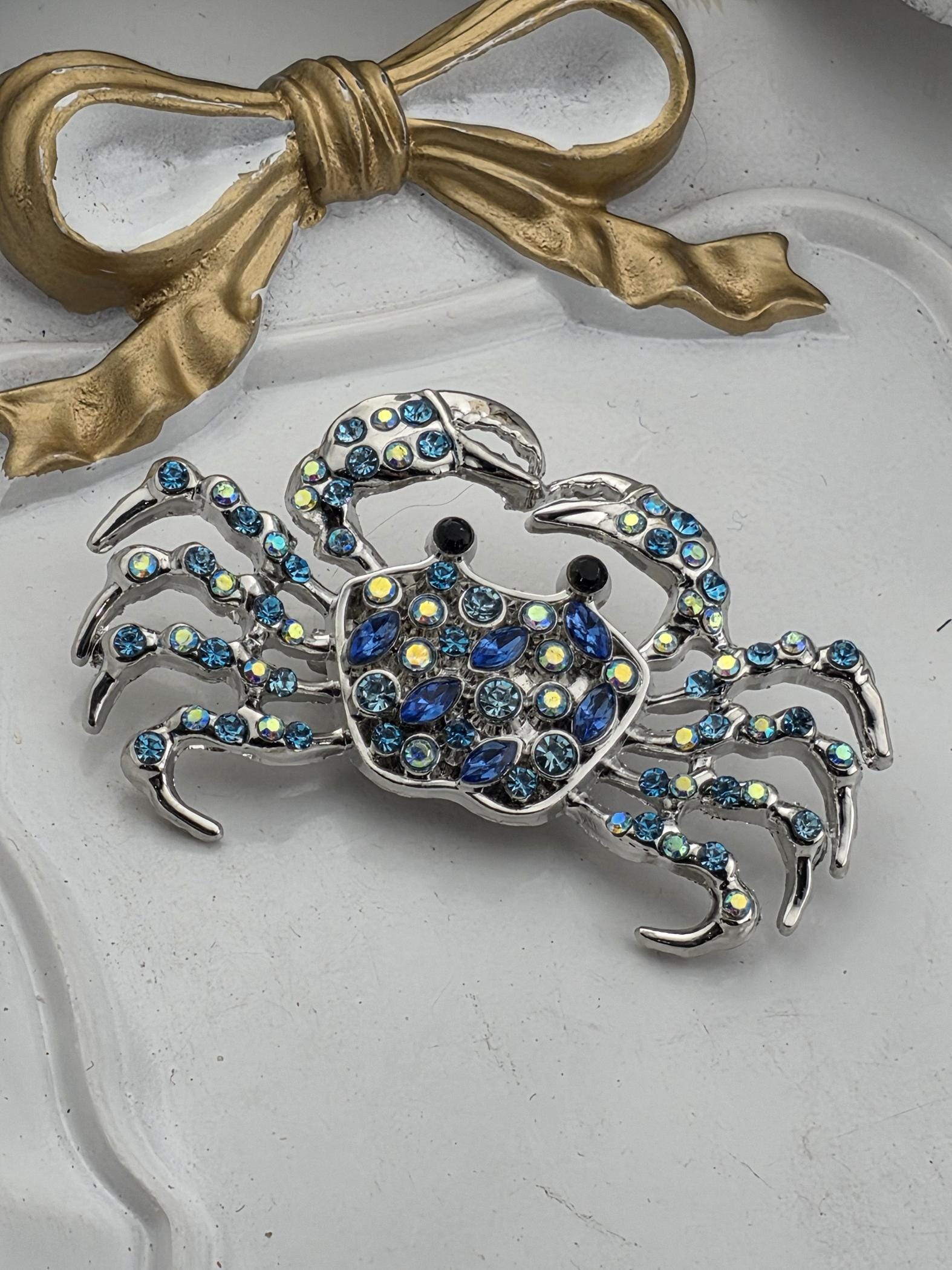 A decorative crab brooch with blue and silver gemstones, placed on a white surface next to a gold ribbon.