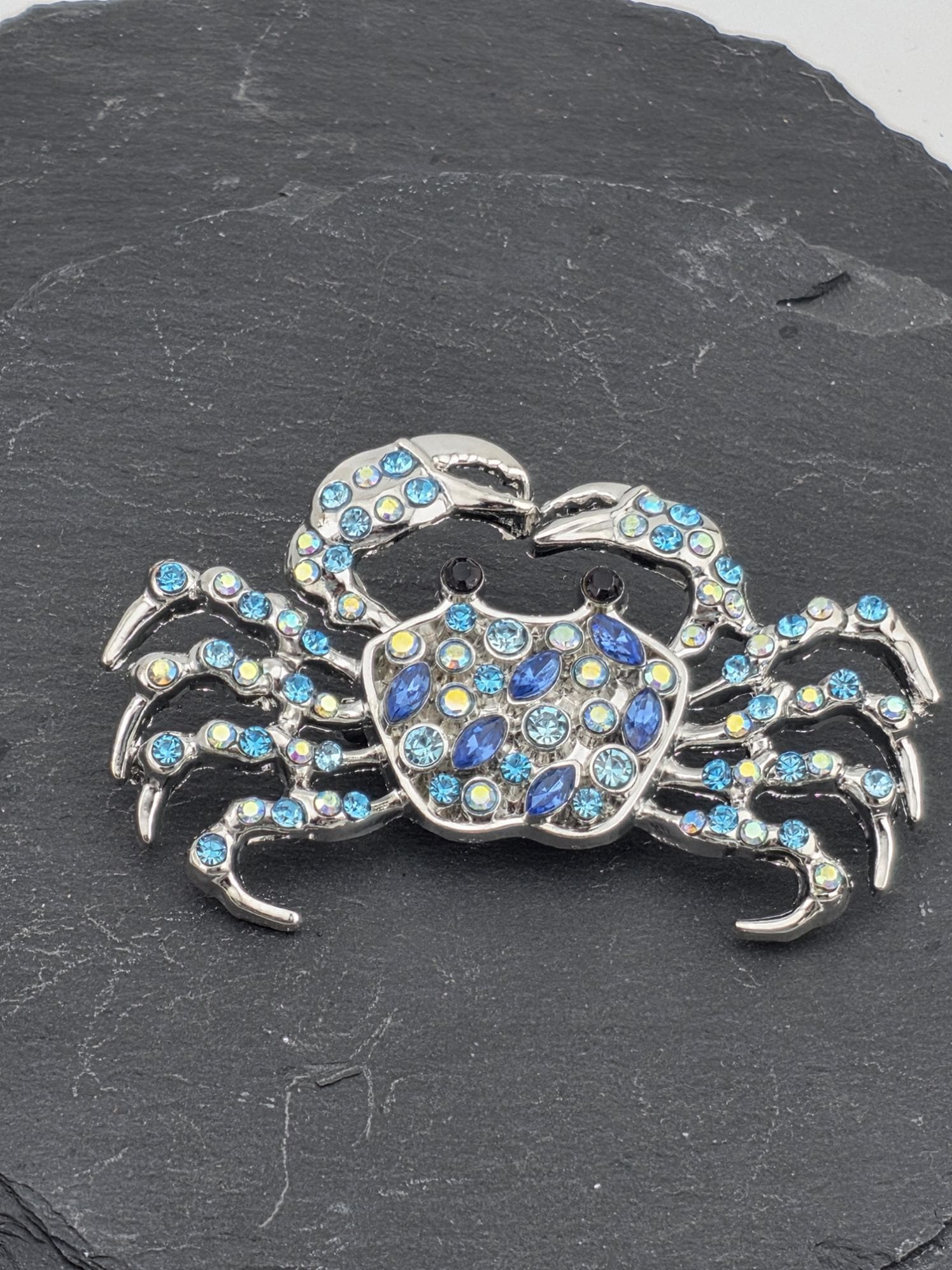 A silver crab brooch adorned with blue and yellow gemstones, resting on a dark surface.