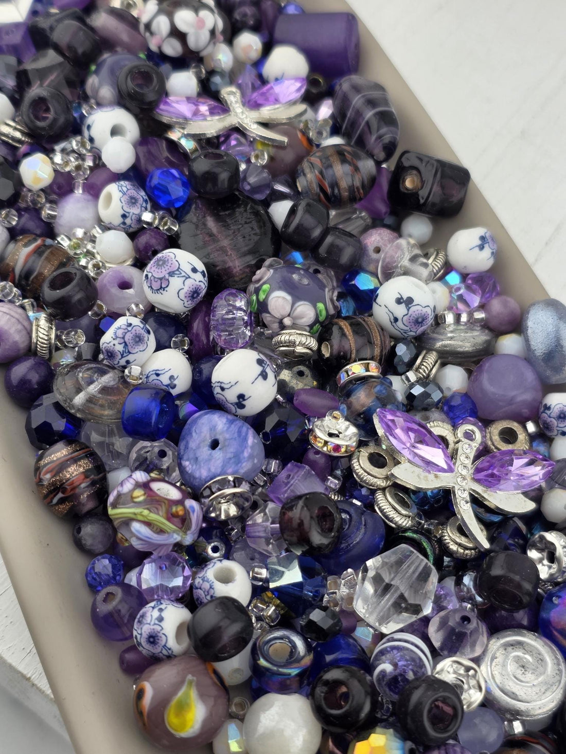 A collection of purple and blue beads of various shapes and sizes. The beads include faceted crystals, round beads with floral patterns, and dark, swirled glass beads. Silver-toned metal accents are interspersed throughout the mix..