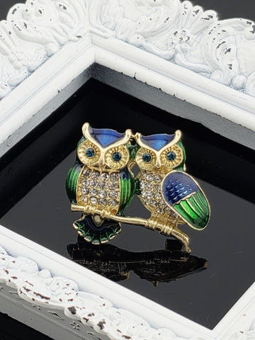A decorative brooch in the shape of two owls, with one owl having a blue body and the other having a green body, is displayed on a reflective surface.