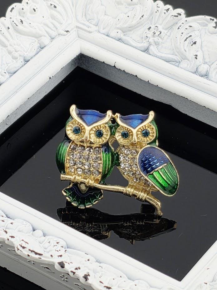 A decorative brooch in the shape of two owls, with one owl having a blue body and the other having a green body, is displayed on a reflective surface.