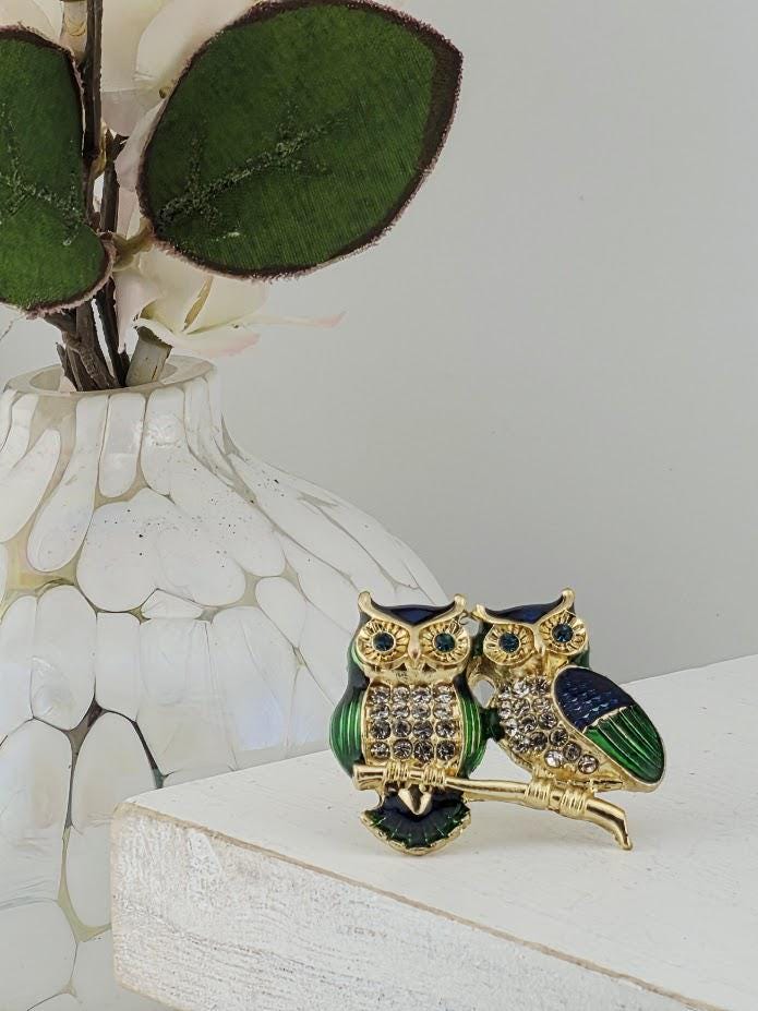 a decorative brooch in the shape of two owls, with one owl being green and the other being blue, both adorned with jewels. The brooch is placed on a white surface, with a white vase containing green and white flowers in the background.