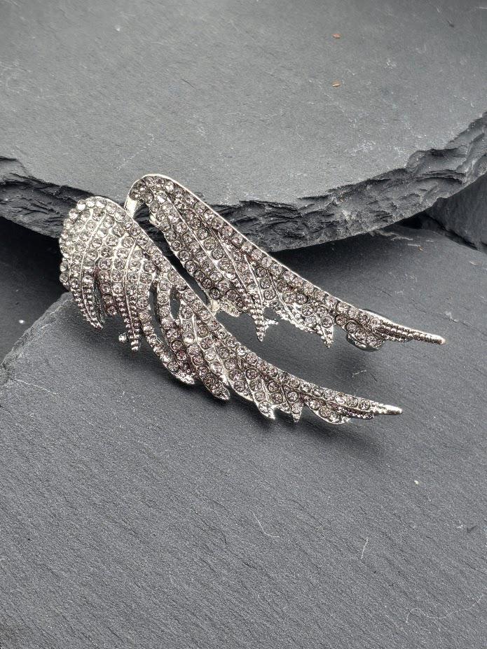 A pair of silver-colored wing-shaped earrings with sparkling rhinestones or crystals on a dark surface.