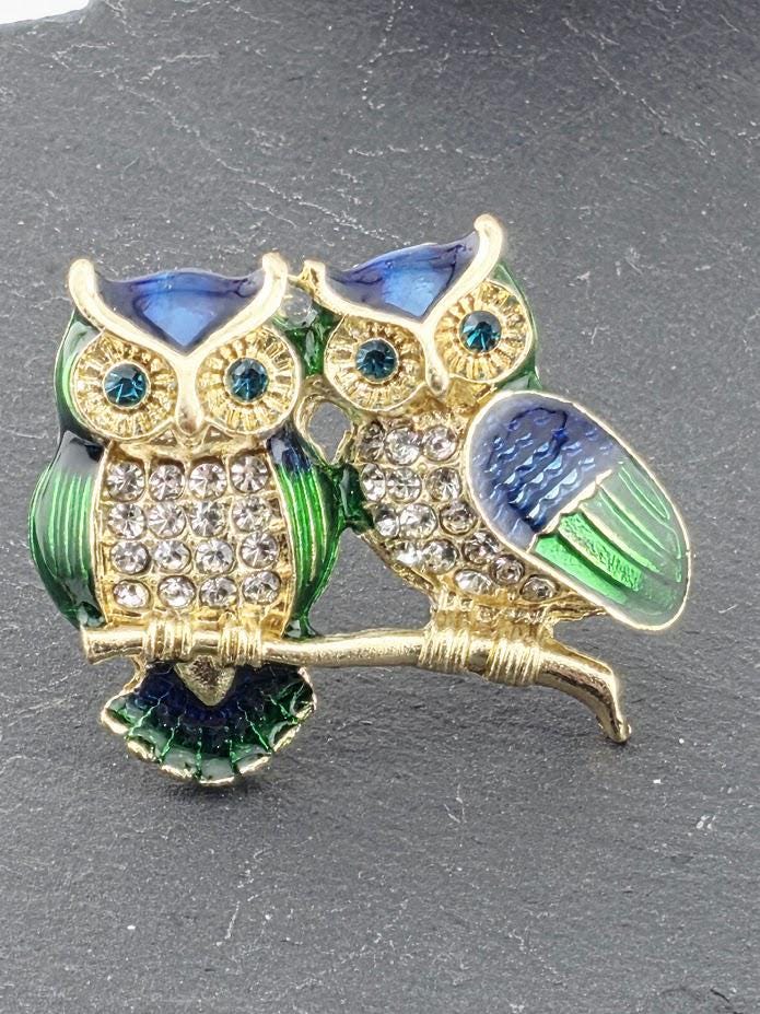 A decorative brooch featuring two ornate owls, one with a blue body and the other with a green body, adorned with gemstones and intricate details.