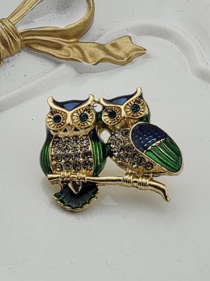 A decorative brooch featuring two ornate owls, one with a green and blue color scheme and the other with a blue and gold color scheme, both adorned with gemstones.