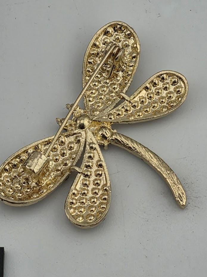 A gold-colored pin in the shape of a dragonfly, with intricate details and a metallic appearance.