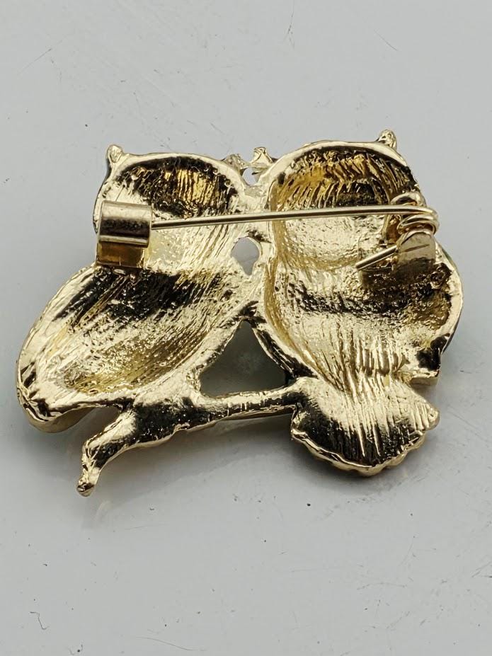 A gold-colored pin in the shape of two golden owls, with intricate details and a shiny finish.