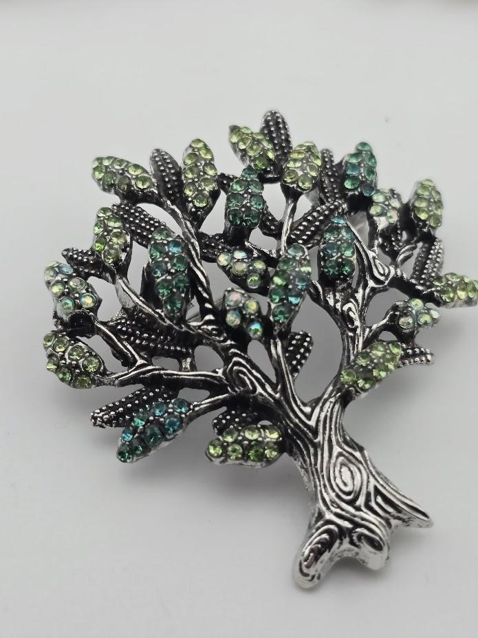 A decorative tree brooch with a silver-colored metal frame and a tree-like design, adorned with green and blue gemstones.