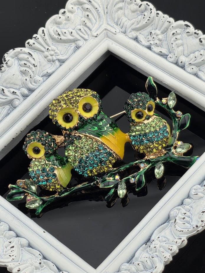 A decorative brooch featuring three ornate owls with green and yellow feathers, set against a black background and framed by an ornate white frame.