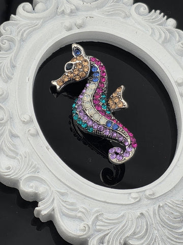 A decorative brooch in the shape of a fish, adorned with colorful gemstones, is displayed within an ornate white frame.