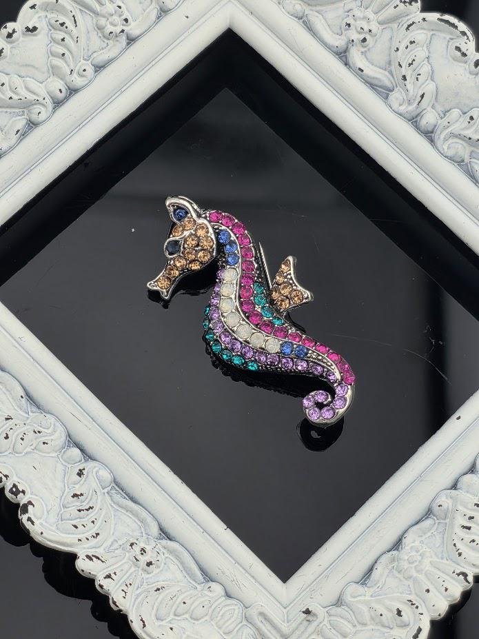 A decorative brooch in the shape of a colorful fish, adorned with gemstones, is displayed within an ornate white frame.