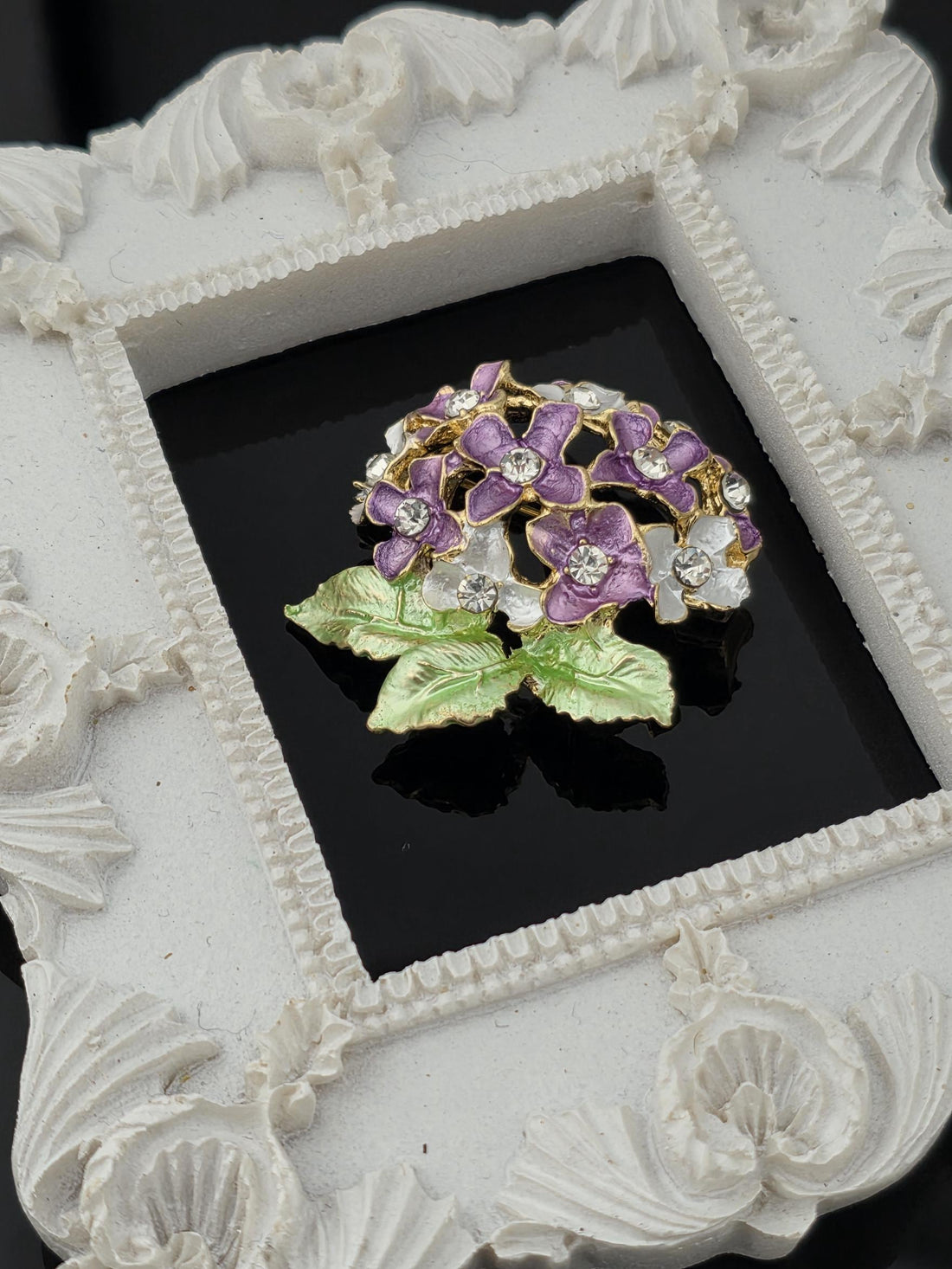 A decorative white frame with a floral design, containing a brooch with purple and green flowers and white stones.
