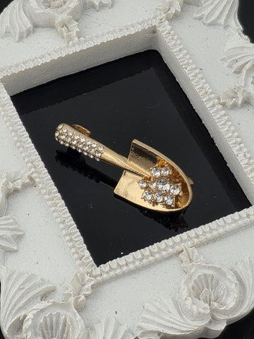 A gold-colored brooch with a shovel-shaped design and small rhinestones is displayed in a white ornate frame.