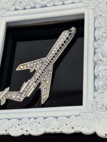 A silver airplane brooch with rhinestones is displayed in a white ornate frame.