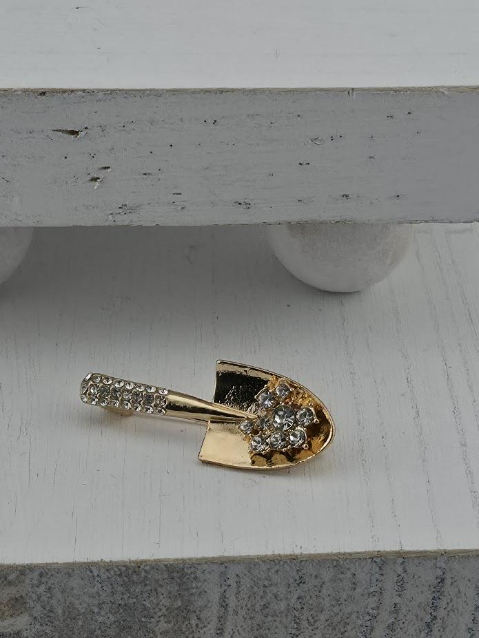 A gold-colored brooch with a diamond-encrusted shovel-shaped design is displayed on a white surface.