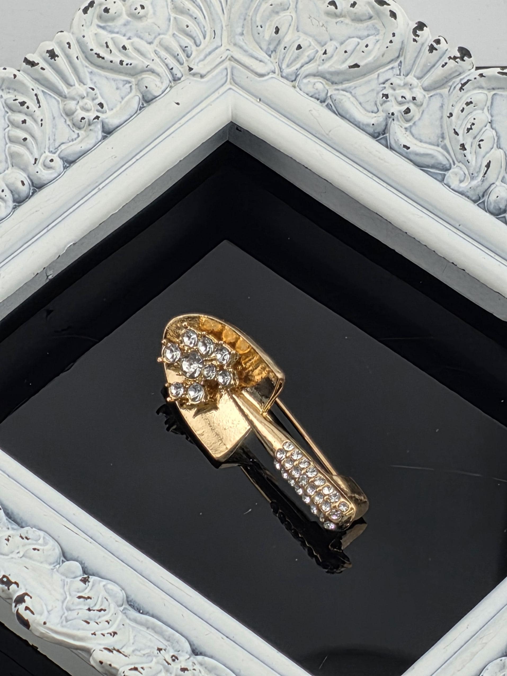 A decorative gold-colored brooch with a floral design and sparkling stones is displayed in a white ornate frame against a black background.