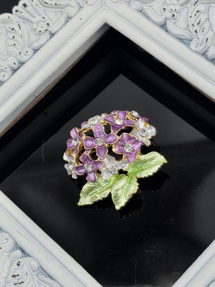 A decorative piece featuring a cluster of purple flowers with green leaves, placed within a white ornate frame against a black background.