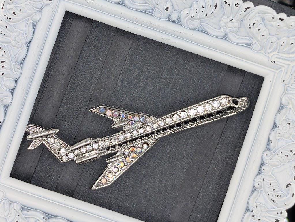 A silver airplane brooch with rhinestones is displayed in a white ornate frame.