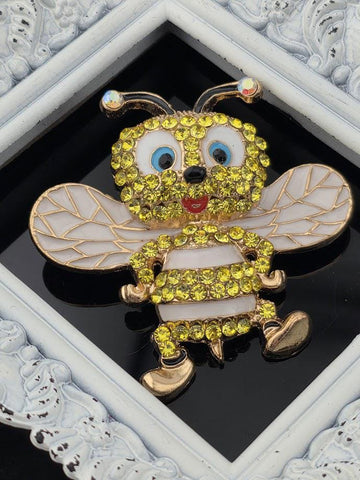 A decorative brooch in the shape of a bee, adorned with yellow and white rhinestones, is displayed against a black background within a white ornate frame.
