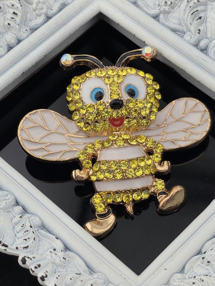 A decorative brooch in the shape of a bee, adorned with yellow and white rhinestones, is displayed against a black background within a white ornate frame.