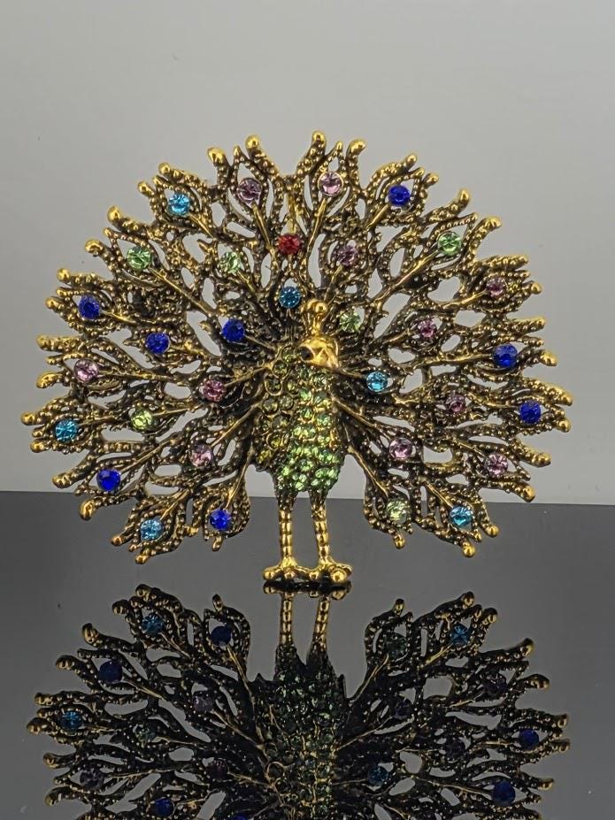 A decorative gold-colored peacock brooch with a colorful array of gemstones, including blue, green, purple, and red stones, set against a gray background.