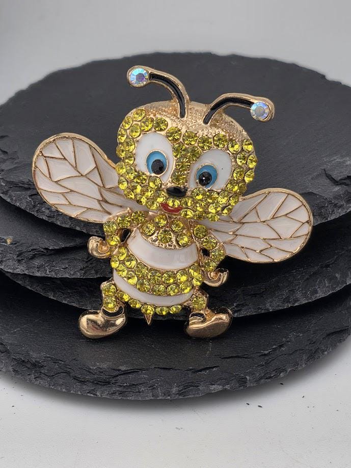 A decorative brooch in the shape of a bee, adorned with rhinestones and gold accents, is displayed on a black slate surface.