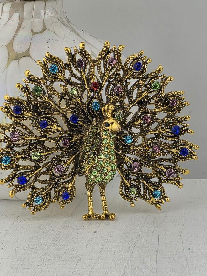 A decorative gold peacock brooch with colorful gemstones, including blue, green, purple, and red stones, is displayed on a white surface.