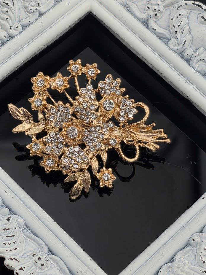 A decorative gold brooch with a floral design and sparkling crystal elements is displayed in a white ornate frame.