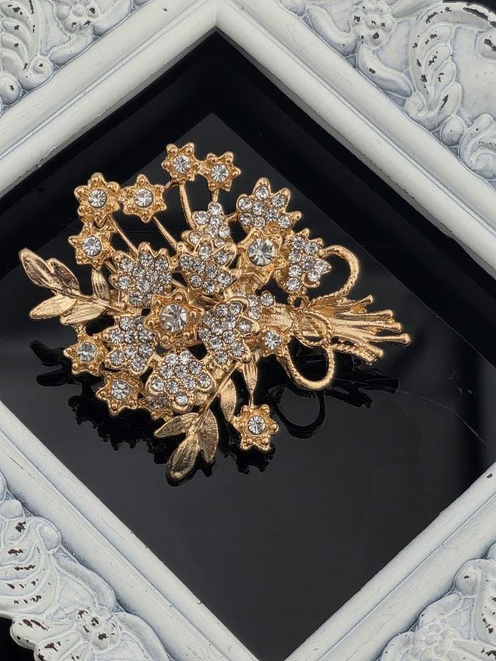 A decorative gold brooch with a floral design and sparkling crystal elements is displayed in a white ornate frame.